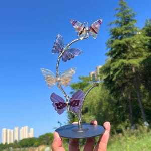 May include: A decorative metal sculpture of four butterflies with translucent wings in shades of purple, blue, and yellow. The butterflies are attached to a silver-coloured branch, mounted on a circular base. The sculpture is displayed outdoors.