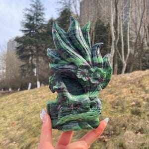 May include: A carved stone sculpture of a fox with a floral and leaf design. The sculpture is green with red and pink accents. The fox is standing on a rock formation. The sculpture is held in a person's hand.