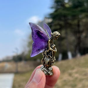 Könnte beinhalten: Kleine, bronzefarbene Feenfigur mit großen, durchscheinenden lila Flügeln. Die Fee sitzt mit den Armen um die Knie. Die Flügel haben ein kreisförmiges Muster. Die Figur wird in einer Hand gehalten.