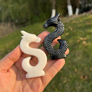 Op de afbeelding: Twee drakenbeeldjes, één wit en één zwart, worden in een hand gehouden. De witte draak is glad, terwijl de zwarte draak een textuur heeft met een schubbenpatroon. Beide bevinden zich in een gebogen, serpentijnachtige pose, met gedetailleerde hoofden.