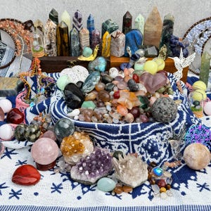 May include: A diverse collection of crystals and gemstones in various forms and colours. The display includes polished spheres, raw clusters, and carved points, arranged on a patterned cloth. The crystals vary from transparent to opaque, with shades of purple, green, and orange.