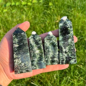 以下が含まれることがあります： さまざまなサイズの4つの緑色のセラフィナイトクリスタルオベリスクが、緑の草を背景に手で持たれています。 オベリスクは独特の羽模様と白い先端を持っています。