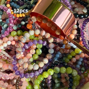 May include: A collection of colorful gemstone beads in various sizes and colors, including purple, green, orange, and clear. A copper-colored scoop is partially visible, and the text "8-12pcs" is in the upper left corner.