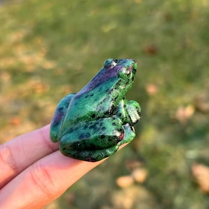 May include: A carved frog figurine in shades of green and purple. The frog has a textured surface with dark spots. The figurine is held up by a finger against a blurred green background.