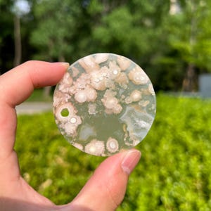 Puede incluir: Una piedra preciosa redonda y pulida con un patrón único de colores verde claro, blanco y beige. La piedra tiene un agujero perforado y se sostiene en una mano sobre un fondo verde borroso.