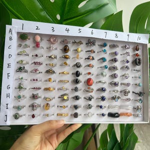 Op de afbeelding: Een display van diverse ringen met verschillende edelstenen en ontwerpen, gerangschikt in een witte tray. De ringen hebben een verscheidenheid aan kleuren en vormen, waaronder smaragden, opalen en andere stenen. De ringen zijn in rijen en kolommen gerangschikt.
