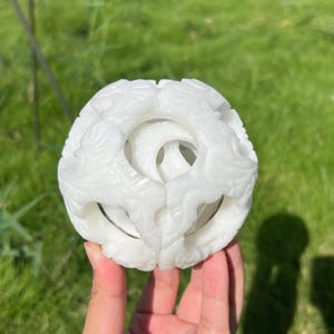 May include: A white, carved sphere with detailed openwork patterns. The sphere is held in a hand against a background of green grass. The carvings have a floral or organic theme, creating a delicate, ornamental piece.