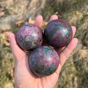 Peut inclure: Trois sphères de pierres précieuses polies, chacune présentant un mélange de teintes rouge profond et vert sarcelle. Les sphères sont tenues dans une main, mettant en valeur leurs formes lisses et arrondies et leurs variations de couleurs naturelles.