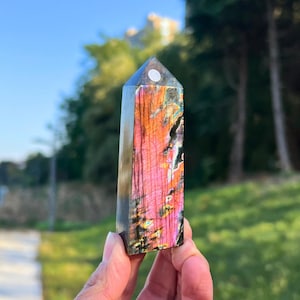 以下が含まれることがあります： 長方形で研磨されたラブラドライトのクリスタルタワー。ピンク、オレンジ、紫の鮮やかな色合いが特徴です。クリスタルは手で持たれており、背景には木々と青い空があります。上部は尖っています。