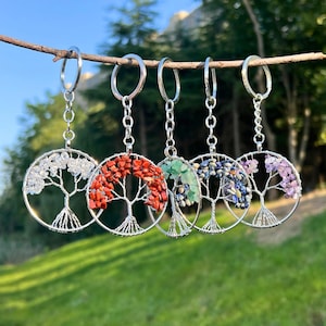 Könnte beinhalten: Fünf Baum des Lebens Schlüsselanhänger mit verschiedenfarbigen Steinen: klar, rot, grün, blau und lila. Jeder Schlüsselanhänger hat ein silberfarbenes Metallbaumdesign und eine silberne Kette und einen Ring zum Befestigen von Schlüsseln.