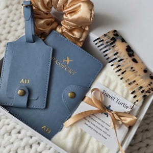 Könnte beinhalten: Geschenkbox mit blauem Reisepasshalter und Gepäckanhänger mit goldenen Initialen, einem goldenen Haargummi, einem Schildpattkamm und einem kleinen weißen Handtuch, das mit einem goldenen Band gebunden ist. Der Reisepasshalter hat eine goldene Flugzeuggrafik.