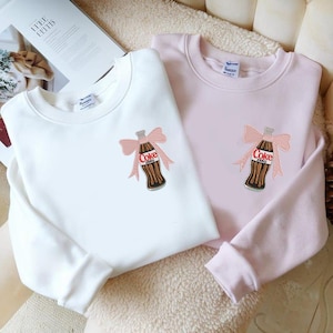 Pode incluir: Duas camisolas, uma branca e outra rosa, são exibidas. Cada uma tem um design de garrafa de Coca-Cola Zero com um laço rosa. As camisolas são feitas de um material macio e são adequadas para uso casual.