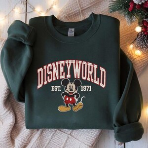 May include: Dark green sweatshirt with the text "DISNEYWORLD" in white and red lettering, with an image of Mickey Mouse. Below the text is "EST. 1971". The sweatshirt is folded on a light-colored surface.