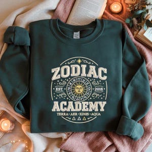 Puede incluir: Sudadera verde oscuro con un gráfico blanco y dorado que dice "ZODIAC ACADEMY" con un sol y símbolos del zodiaco. La sudadera tiene cuello redondo y mangas largas. El texto "EST. 2018" también está incluido.