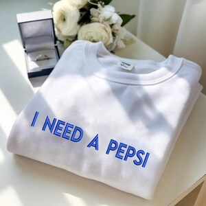 Puede incluir: Sudadera blanca con el texto azul bordado "I NEED A PEPSI". La sudadera doblada está sobre una superficie blanca, con un joyero y un ramo de flores blancas en el fondo.