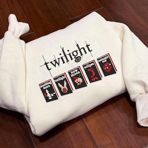 Puede incluir: Sudadera crema con la palabra "twilight" bordada en negro. Debajo, cinco pequeñas portadas de libros bordadas con títulos como "Breaking Dawn" y "Midnight Sun". La sudadera tiene puños acanalados y cuello redondo.