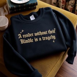 Könnte beinhalten: Ein marineblaues Sweatshirt mit goldenem Text, der lautet: „Ein Leser ohne seinen Kindle ist eine Tragödie“. Das Sweatshirt liegt auf einem gelben Kissen. Eine Tasse, Bücher und ein Teller sind im Hintergrund.