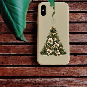 Pode incluir: Uma capa de telefone bege com um design de &aacute;rvore de Natal. A &aacute;rvore &eacute; decorada com flores brancas, enfeites dourados e um la&ccedil;o verde. A capa est&aacute; colocada sobre um fundo de madeira com uma folha verde no canto superior esquerdo.