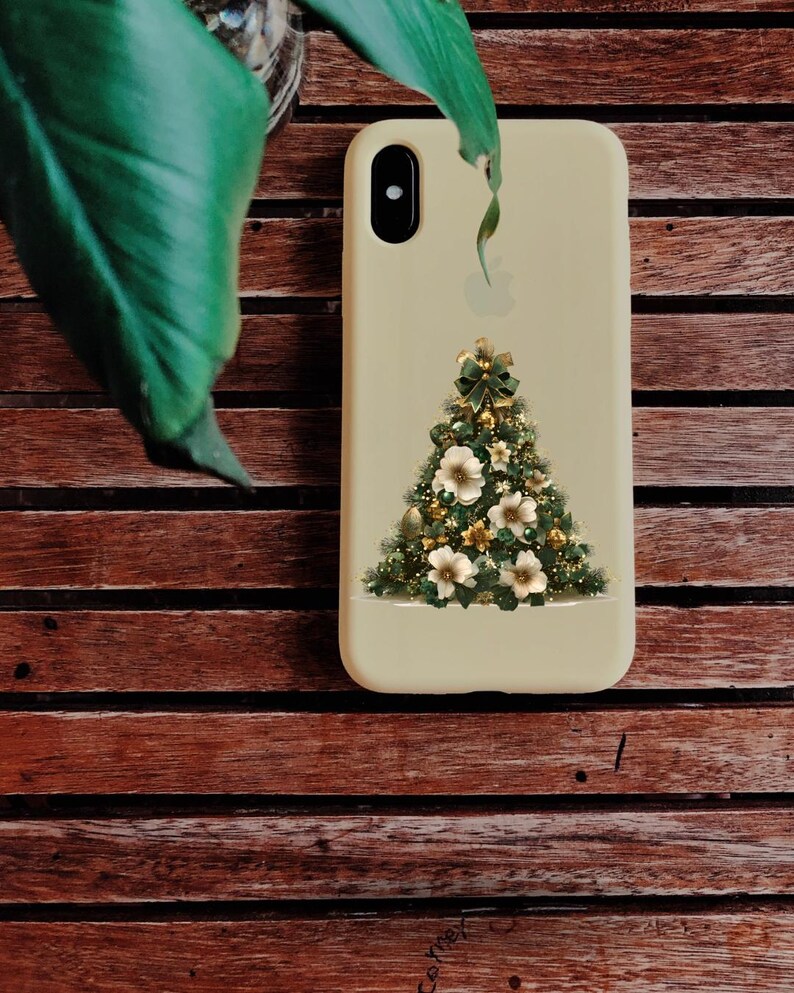Pode incluir: Uma capa de telefone bege com um design de &aacute;rvore de Natal. A &aacute;rvore &eacute; decorada com flores brancas, enfeites dourados e um la&ccedil;o verde. A capa est&aacute; colocada sobre um fundo de madeira com uma folha verde no canto superior esquerdo.