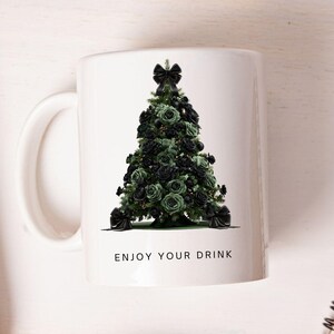 Pode incluir: Caneca de cer&acirc;mica branca com um desenho de &aacute;rvore de Natal feito de rosas pretas e verdes, adornada com la&ccedil;os pretos. A caneca tem uma al&ccedil;a branca e o texto "ENJOY YOUR DRINK". Uma caneca festiva com tema de feriado.