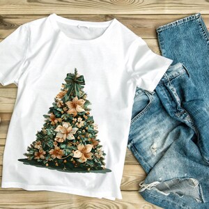 Pode incluir: Camiseta branca com um desenho de &aacute;rvore de Natal. A &aacute;rvore &eacute; decorada com folhagem verde, flores bege e um la&ccedil;o verde. Jeans azuis est&atilde;o ao lado da camiseta em uma superf&iacute;cie de madeira.