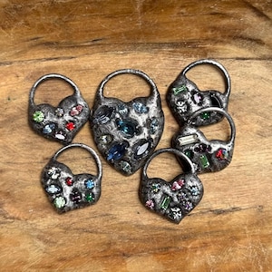 May include: A collection of five heart-shaped pendants with a dark, textured metallic finish. Each pendant features an integrated circular loop at the top and is embedded with various small, multi-colored gemstones in red, blue, green, and clear.