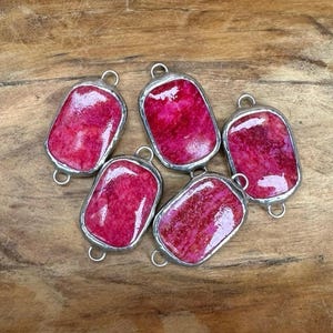 May include: Five rectangular pendants with a vibrant pink, textured stone set in a silver-colored metal frame. Each pendant has a small loop at the top for attaching to a chain or other jewelry component. The pendants are arranged on a wooden surface.
