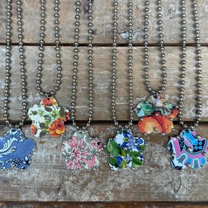 May include: Six necklaces with flower-shaped pendants. Each pendant features a unique floral or patterned design in various colors, including blue, pink, orange, and green. The necklaces have silver-colored ball chains, displayed on a rustic wooden surface.