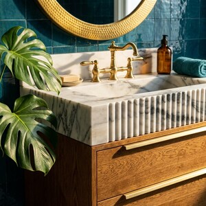 May include: A white marble bathroom sink with a fluted front design, featuring a brass faucet, a wooden vanity cabinet with two drawers, a round gold-rimmed mirror, a brown soap dispenser, and a teal towel, accented by a large green monstera leaf.