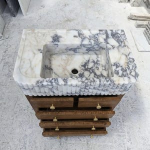 May include: A bathroom vanity featuring a white and grey marble countertop with a rectangular basin and a fluted front edge. The base is constructed from brown wood with four horizontal drawers, each equipped with two small, round, gold-toned knobs.
