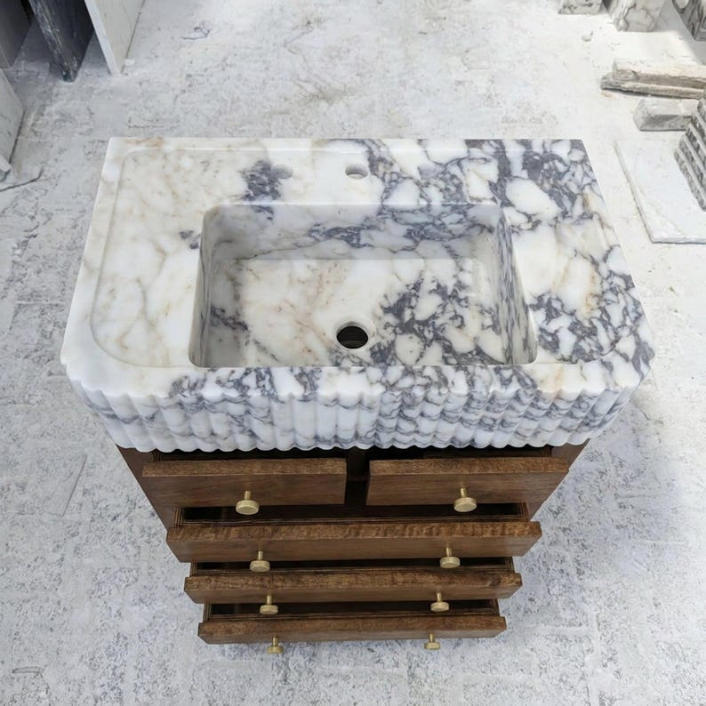 May include: A bathroom vanity featuring a white and grey marble countertop with a rectangular basin and a fluted front edge. The base is constructed from brown wood with four horizontal drawers, each equipped with two small, round, gold-toned knobs.