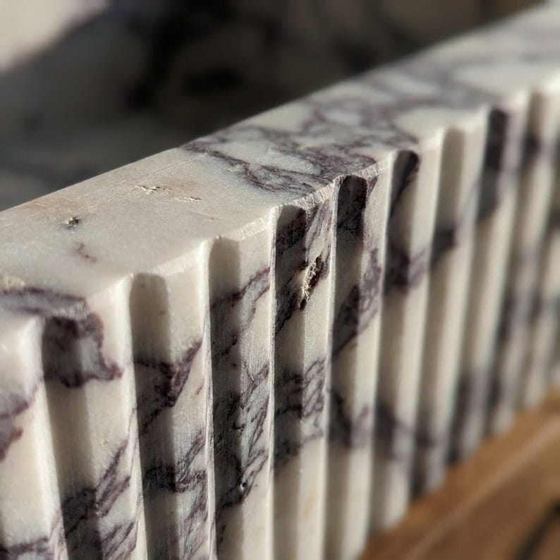 May include: A white marble basin featuring a distinct fluted or ribbed exterior design with prominent dark purple veining throughout the stone surface. The object displays a series of vertical grooves carved into the thick, textured marble edge.