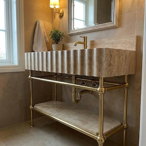 Può includere: Un mobile da bagno con lavabo in pietra travertino beige scanalato, sostenuto da una struttura in tubi di ottone lucido. L'unità include un ripiano inferiore in pietra abbinato, un rubinetto in ottone e tubature a vista sotto il lavabo.