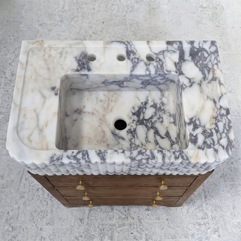 May include: A rectangular bathroom vanity featuring a white and grey marble countertop with a scalloped front edge and a central basin. The base is constructed from brown wood and includes three horizontal drawers with small round brass-toned knobs.