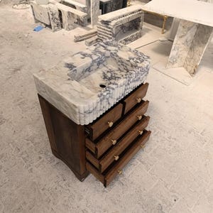 May include: A bathroom vanity featuring a white and grey veined marble sink with a fluted exterior design. The base is constructed from dark brown wood and includes four stacked drawers, each equipped with a small, round, gold-toned metal knob.