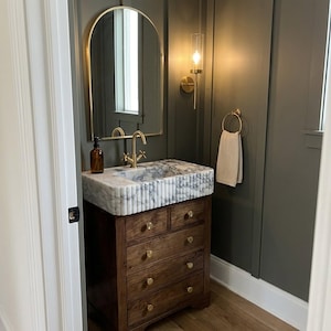 May include: A bathroom vanity featuring a dark wood cabinet with five drawers and brass knobs, topped with a fluted white marble sink. A gold arched mirror, a gold faucet, a wall-mounted light fixture, and a gold towel ring with a beige towel.