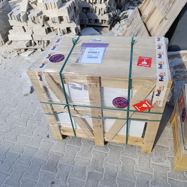 May include: A rectangular wooden shipping crate reinforced with diagonal support beams, wrapped in clear plastic film and secured with green plastic straps. The crate features FedEx branded adhesive tape, fragile warning labels, and shipping documents.