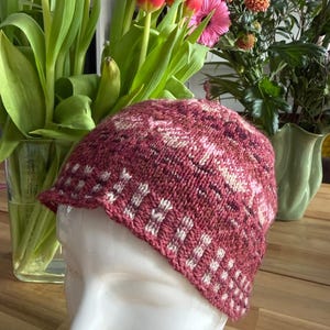 Op de afbeelding: Een gebreide cloche hoed in bordeaux en wit, getoond op een mannequin hoofd. De hoed heeft een patroon met witte geometrische vormen. De achtergrond bevat een vaas met tulpen en andere bloemen.