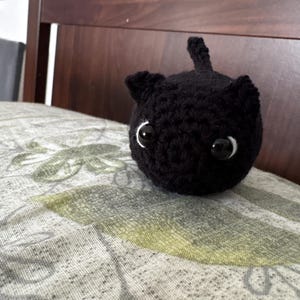 Puede incluir: Un pequeño gato negro tejido a ganchillo con ojos ribeteados en blanco y una pequeña cola. El gato hecho a mano está sentado sobre una almohada estampada con diseños florales verdes y grises. Las orejas del gato son puntiagudas.