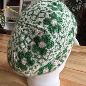 Op de afbeelding: Een groen en beige gebreide baret met een bloemmotief. De baret wordt getoond op een witte mannequinhoofd, waarbij het ingewikkelde ontwerp en de textuur van het breiwerk worden getoond. Het kleurenpalet van de baret omvat tinten groen en beige.