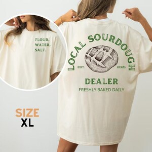 Op de afbeelding: Crèmekleurig XL-shirt met groene tekst en afbeeldingen. De voorkant toont "FLOUR. WATER. SALT." De achterkant heeft een broodafbeelding met "LOCAL SOURDOUGH DEALER" en "FRESHLY BAKED DAILY".