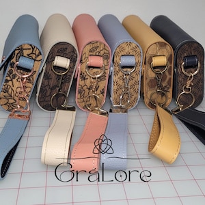 May include: A collection of leather cases in various colors, including blue, cream, pink, and tan. Each case features a wooden panel with engraved floral or honeycomb designs and a matching wrist strap. The cases are designed for carrying small items.