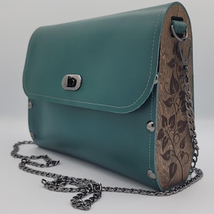 Puede incluir: Bolso de mano de cuero verde azulado con un panel lateral de madera con un diseño de hojas. El bolso tiene una correa de cadena plateada y un cierre de broche plateado. El bolso es rectangular con bordes redondeados.