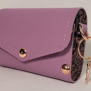 May include: A lavender leather key holder with a flap closure and gold-colored rivets. The key holder has a decorative floral pattern on the side and a gold-colored key ring and clasp.