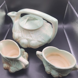 May include: A vintage ceramic tea set in a light green color, featuring a teapot with a lid and two matching creamers. The set has a textured, cabbage-like design, with a light beige interior.