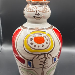 May include: A whimsical ceramic bottle with a painted face and a small orange hat. The bottle features a white base with red, yellow, and brown accents, and a design of a person holding a flower. A unique decorative piece.