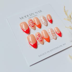 May include: A set of ten artificial nails in various designs. The nails feature a peach base with red tips, orange accents, and floral patterns. The card reads "MODERN NAIL DESIGNS" and "BASED IN BROOKLYN, NY."