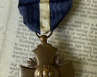 May include: A bronze military medal with a blue and white striped ribbon. The medal features a cross design with a central emblem of a ship. The background is an aged paper.