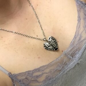 Puede incluir: Collar plateado con un colgante en forma de corazón. El colgante tiene un diseño detallado y texturizado. El collar se lleva sobre una camiseta gris con ribete de encaje. La cadena es delicada.