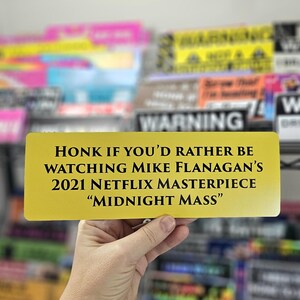 Peut inclure: Un panneau de couleur or avec du texte noir qui dit "HONK IF YOU'D RATHER BE WATCHING MIKE FLANAGAN'S 2021 NETFLIX MASTERPIECE "MIDNIGHT MASS""